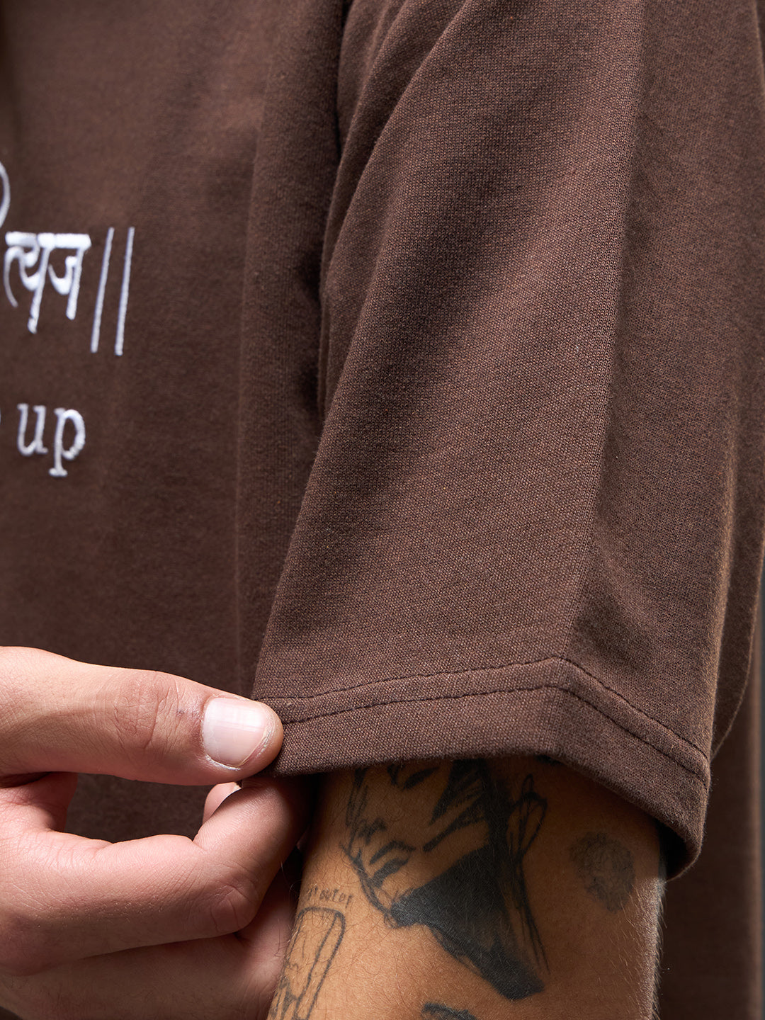 Brown recycled oversized tee by Rudrajap with "Never Give Up" embroidery, shown flat with visible round neckline and relaxed fit.